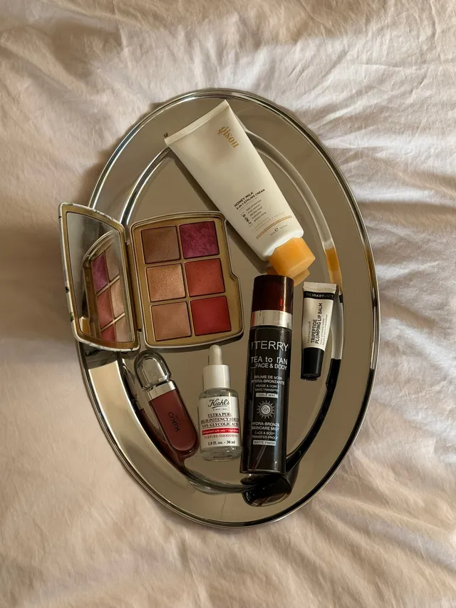 Make-up and skincare — a blusher palette, serum, lip products and a tanning mist — laid out on an oval silver tray on crumpled linen
