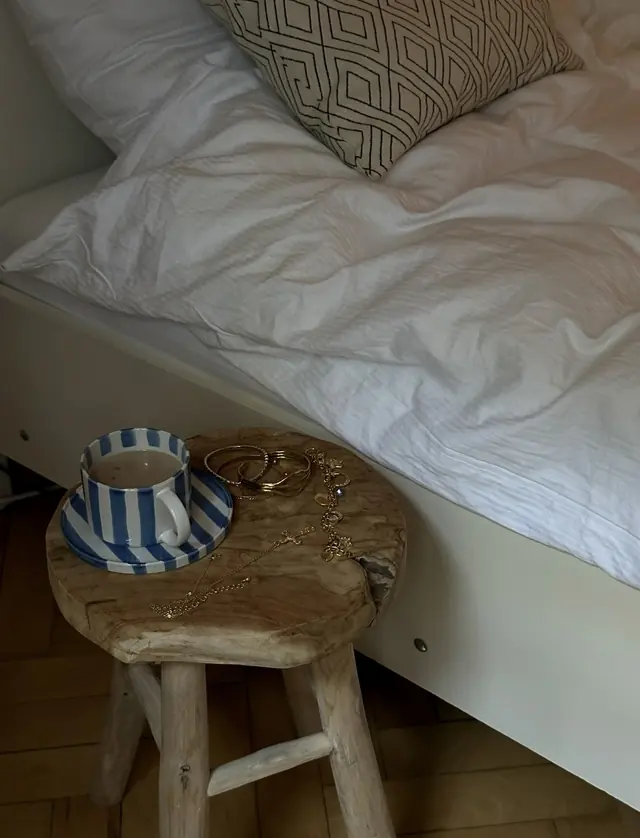 A striped cup and saucer and gold jewellery on a wooden stool beside a white bed