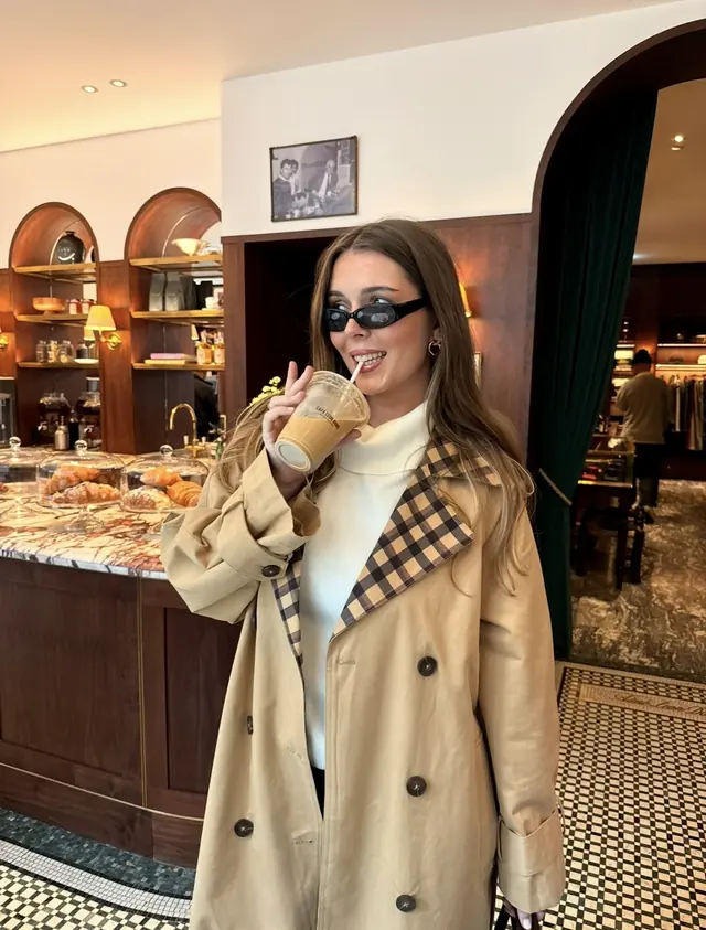 A woman in a checked-lapel trench coat drinking an iced coffee in a wood-panelled café