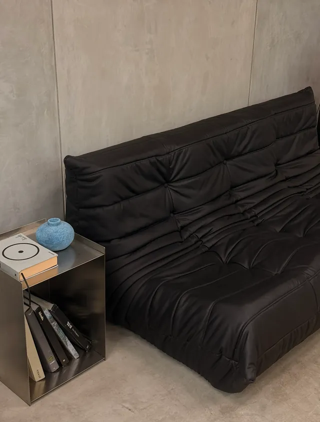 A low black leather sofa against a poured-concrete wall, with books and a blue vase on a steel side table
