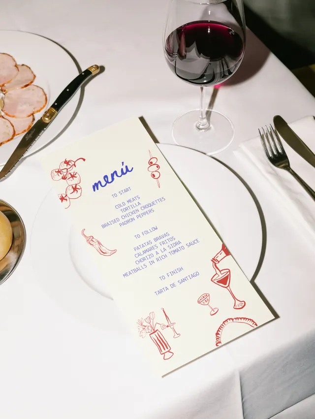 An illustrated menu card, hand-drawn in red and blue, on a white-clothed table laid with wine, bread and charcuterie
