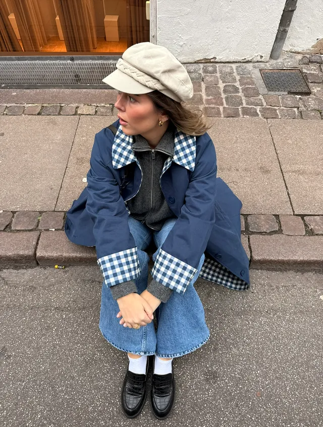 A woman in a navy gingham-lined trench coat and corduroy baker boy cap sitting on a cobbled kerb
