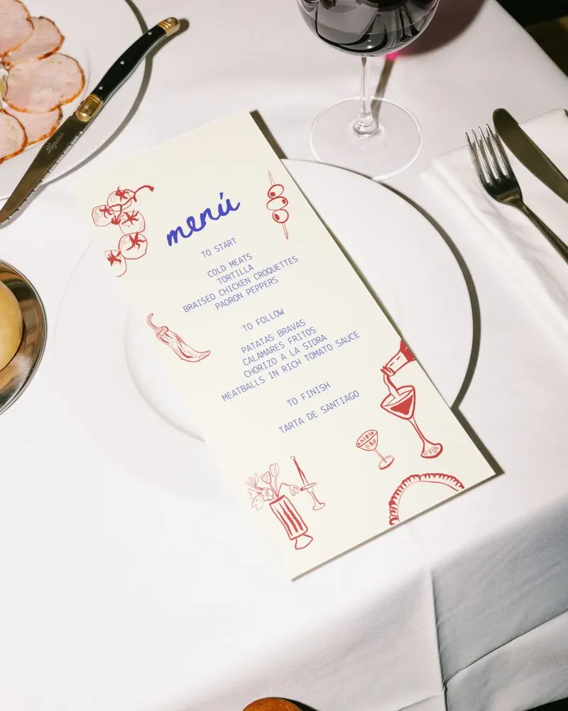 A printed menu decorated with the Apéritif illustrations, on a set table beside a glass of red wine